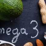Assortment of omega-3 rich foods including salmon, whole fish, avocado, seeds, nuts, and oil, arranged on a dark surface with the words “Omega 3” written in the center.