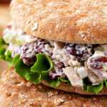 Whole wheat sandwich filled with creamy chicken salad, red grapes, and lettuce, served with celery sticks on a wooden board.