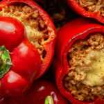 Overhead view of red bell peppers stuffed with a savory chili-style filling and melted cheese, surrounded by fresh vegetables and sauce on a rustic table.