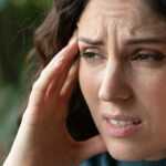 Woman holding her temples with a worried expression, appearing stressed or experiencing a headache while sitting indoors near a window.