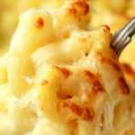 Close-up of creamy baked macaroni and cheese with golden melted cheddar, being lifted on a spoon, with visible stretchy cheese and parsley garnish.