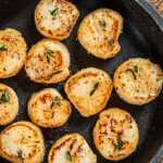 Seared sea scallops with a golden crust served in a black skillet, garnished with herbs and accompanied by garlic cloves and a bowl of coarse salt on a rustic wooden board.