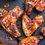 Grilled barbecue chicken wings with charred glaze sit on a wooden cutting board beside a small copper pot of sauce and a carving knife, with sauce drizzled on the board.