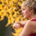 Woman practicing Tai Chi QiGong outdoors by a river, performing a slow, flowing arm movement surrounded by autumn foliage.