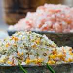 Bowls of gourmet sea salts, including herbal seasoning salt, Himalayan pink salt, and coarse white salt, displayed with fresh herbs on a rustic wooden table.