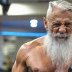 Fit senior man with a long white beard lifting a dumbbell in a gym, performing a bicep curl with visible muscle definition and focused expression.