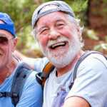 Two cheerful senior men wearing casual hiking clothes and backpacks sit together on a forest trail, smiling and laughing while taking a break outdoors.
