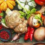 Overhead view of fresh vegetables and ingredients including pumpkin, celery, mushrooms, herbs, and spices arranged around a cutting board with a halved pumpkin and seeds.