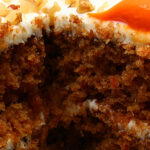 Slice of moist carrot cake with cream cheese frosting, decorated with icing carrots and chopped nuts, with a fork taking a bite from the soft layered cake.