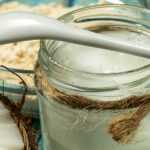 Glass jar of coconut oil with a white spoon, surrounded by fresh coconut halves and pieces on a turquoise wooden surface.