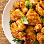 A bowl of crispy Asian citrus chicken coated in a glossy orange sauce, garnished with fresh parsley, served on a rustic wooden board with chopsticks.