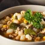 Bowl of cozy chicken and white bean soup with vegetables, topped with fresh parsley, served on a rustic wooden table with a spoon nearby.