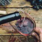 A hand pours red wine from a bottle into a glass on a rustic wooden table surrounded by fresh black grapes, herbs, and a corkscrew.