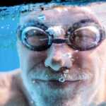 Smiling senior man wearing swim goggles underwater in a pool giving two thumbs up, illustrating the health benefits of swimming and active aging.