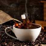 Steaming cup of black coffee with sugar cubes splashing in, surrounded by scattered coffee beans, a burlap sack, and a vintage wooden coffee grinder on a rustic wooden table.