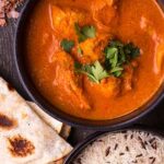 Bowl of creamy chicken curry garnished with fresh cilantro, served with basmati rice and warm naan bread on a dark wooden table, with herbs and spices scattered around.