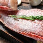 Two raw fish fillets on a wooden board, garnished with sprigs of rosemary, next to garlic cloves and a bowl of pink Himalayan salt.