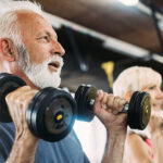 A senior man and woman lifting dumbbells while seated on workout benches in a gym with various exercise equipment in the background.