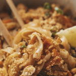 Bowl of vegan Pad Thai with rice noodles, crushed peanuts, green onions, and a lime wedge, served with chopsticks on a wooden table.