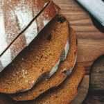 Sliced loaf of dark rye sourdough bread on a wooden cutting board with a knife, set on a rustic table with a cloth backdrop.