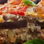 Slice of traditional Greek moussaka layered with eggplant, minced meat, and creamy béchamel sauce, garnished with fresh basil and served on a rustic plate with tomatoes in the background.