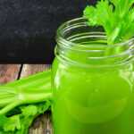 Fresh celery juice in a mason jar garnished with celery leaves, with celery stalks and lime slices on a rustic wooden table.