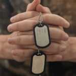 A soldier in camouflage uniform sits with hands clasped, holding a pair of military dog tags, appearing contemplative in soft, warm light.