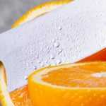 Close-up side view of a chef’s knife slicing a fresh orange in half on a wooden surface, with whole and cut oranges visible in the background.
