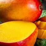 Fresh ripe mangoes on a wooden surface, including a whole mango, a halved mango, and neatly cubed mango pieces, surrounded by green leaves.