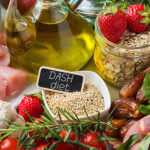 An assortment of DASH diet foods including fresh fruits, vegetables, salmon, chicken, eggs, olive oil, whole grains, nuts, and quinoa arranged on a table.