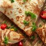 Golden-brown quesadilla wedges arranged on a wooden board, garnished with chopped cilantro and sliced red chilies, served alongside a bowl of creamy cilantro yogurt sauce with lime wedges on a rustic table.