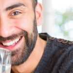 Smiling man holding and drinking a glass of water indoors, with natural light and a bright, clean background.