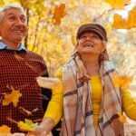 Happy older couple enjoying autumn in a forest, tossing colorful fallen leaves in the air and smiling among golden foliage.