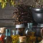 Rustic still life of dried medicinal herbs hanging above a wooden table with glass bottles of infused oils, a mortar and pestle, and bundles of plants arranged in a natural apothecary setting.