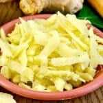 Fresh ginger root with sliced pieces and grated ginger in a small bowl, alongside ground ginger in a wooden spoon on a rustic wooden table.