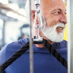 Older man with gray hair and beard using a cable machine for a chest exercise at the gym.